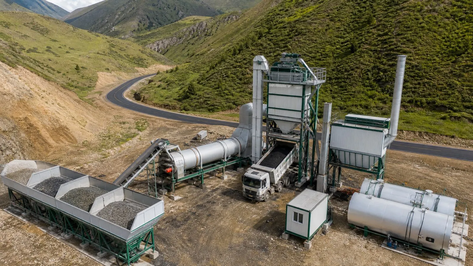 Vista aérea de una planta de asfalto en contenedores junto a una carretera nueva entre cerros verdes (imagen ilustrativa)