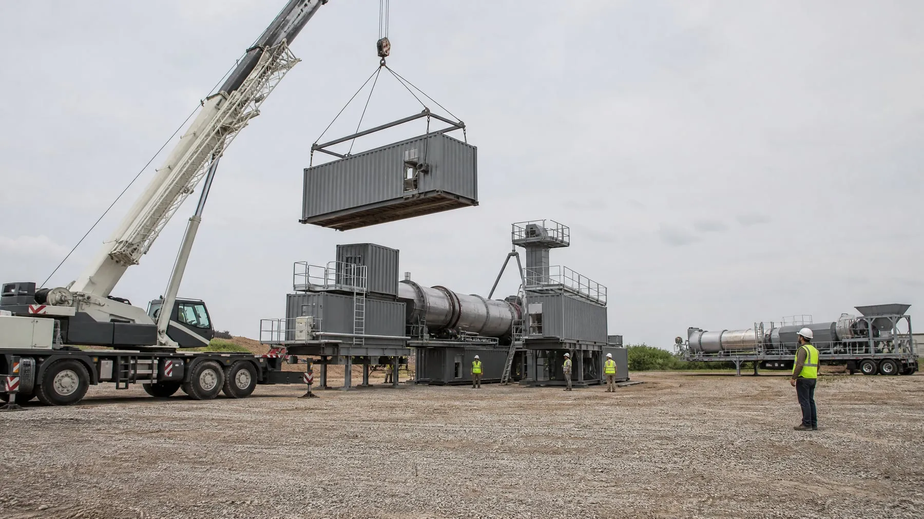 Crane lifting a container-sized asphalt plant module into place, with a trailer-mounted plant in the background (illustrative image)